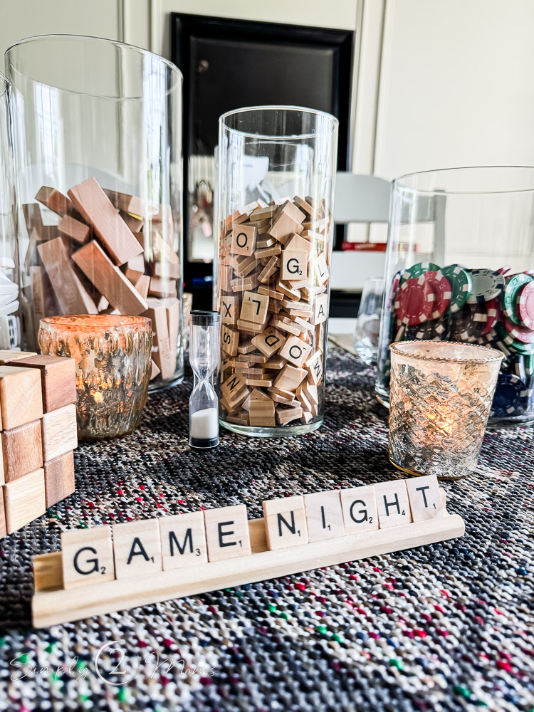 A table is set up for a game night, featuring glass jars filled with Jenga blocks, Scrabble tiles, and poker chips. Small lit candles and an hourglass add to the ambiance. Scrabble tiles on a rack spell out "GAME NIGHT." A textured, colorful table runner enhances the setup.