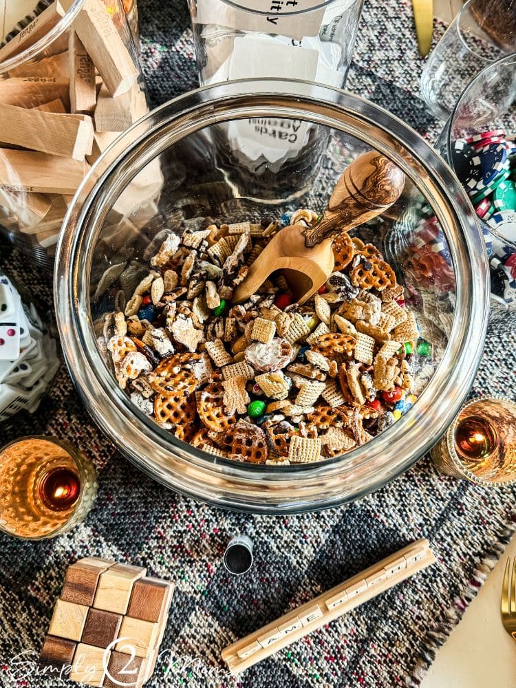 A glass bowl filled with a mixture of snacks, including pretzels, cereal, chocolates, and nuts, is placed on a patterned cloth. A wooden scoop rests inside the bowl. Surrounding items include candles, playing cards, a block tower game, and a wooden puzzle—recipes for a winning game night party.