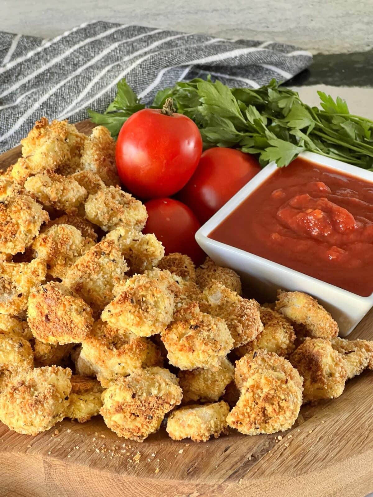 A wooden board filled with crispy, breaded cauliflower bites next to a small square bowl of marinara sauce, garnished with fresh parsley and tomatoes in the background. Perfect for your recipes for a winning game night party, a striped napkin is partially visible on the left side.