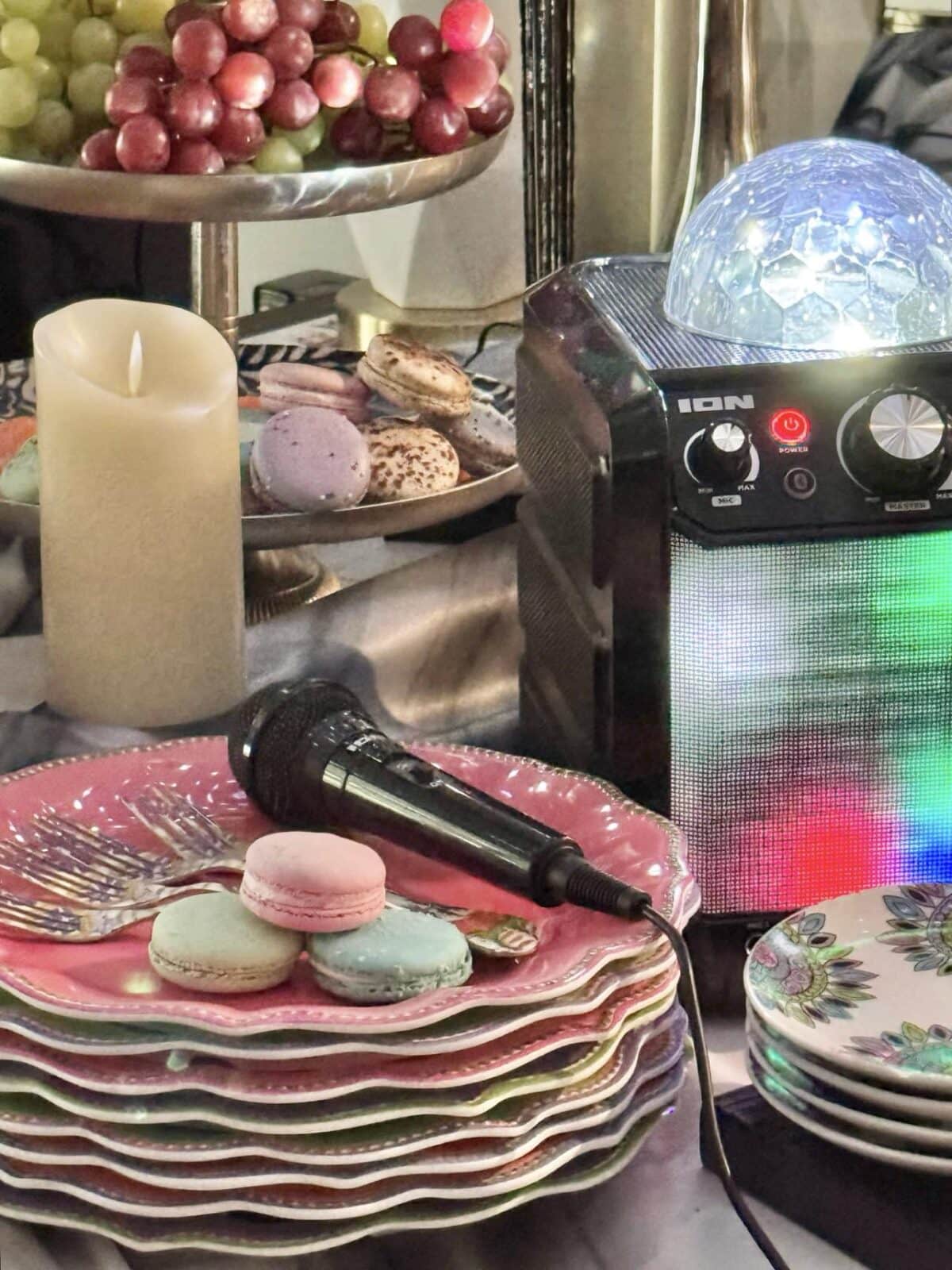 A table setup with a stack of colorful plates, macarons on top, and a microphone resting beside them. There's a faux candle, a bowl of grapes, and a box-shaped speaker or lighting device with a dome light in the background.