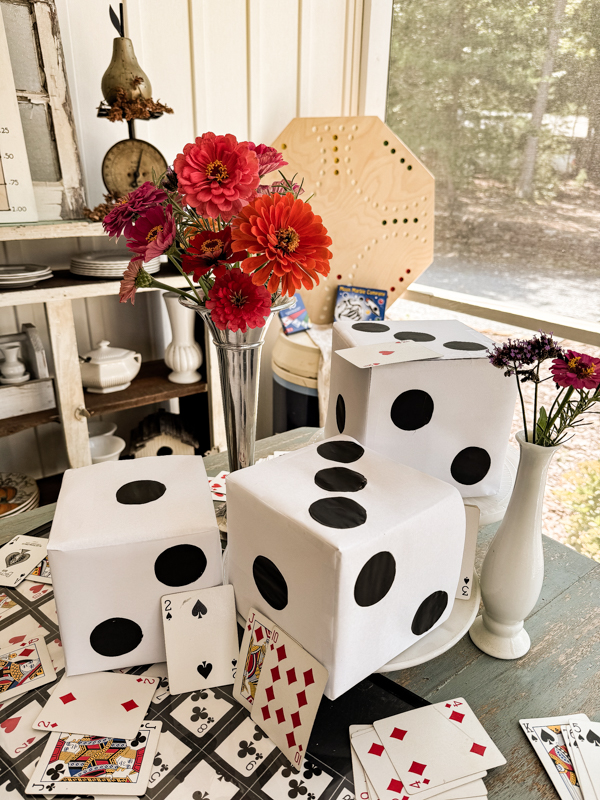 A table is decorated with large dice, scattered playing cards, and vases of colorful flowers. The background features additional decor including a clock, a large wooden cheese board, and a window with a view outside.