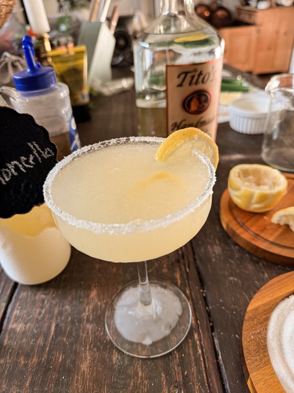 A lemon cocktail with a sugared rim and a lemon slice garnish is placed on a wooden table. Surrounding the drink are a bottle of Tito's Vodka, a half-squeezed lemon, a small bottle of homemade lemonade, and various bar accessories – perfect for recipes for a winning game night party.