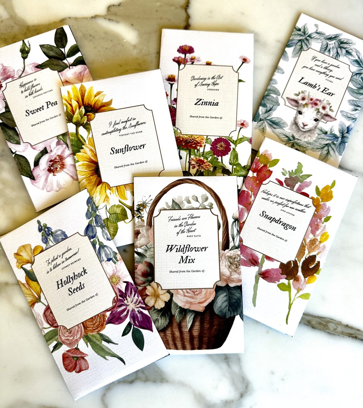 A collection of flower seed packets is spread out on a marble surface. The packets include various flowers such as Sweet Pea, Sunflower, Zinnia, Lamb's Ear, Wildflower Mix, and Snapdragon. Among them are Harvest Store and Share Hollyhock Seeds, each illustrated with vibrant floral artwork.