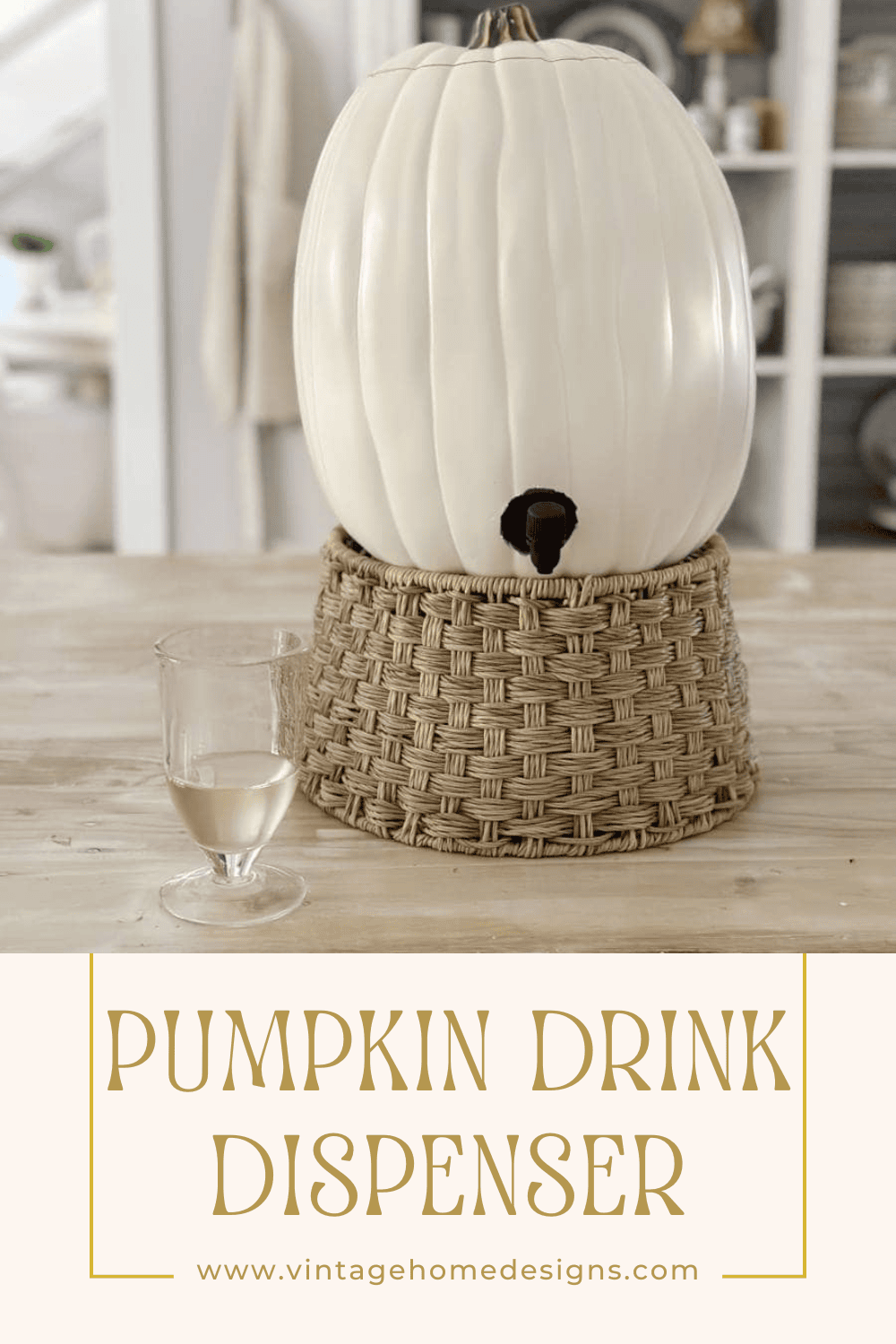 A white pumpkin drink dispenser is placed atop a woven basket on a light wooden table, doubling as a stylish pumpkin wine holder. Beside it is a glass of white wine. In the background, there's a kitchen setting with shelves and various decor items. Text at the bottom reads, "Pumpkin Drink Dispenser" and a website URL.