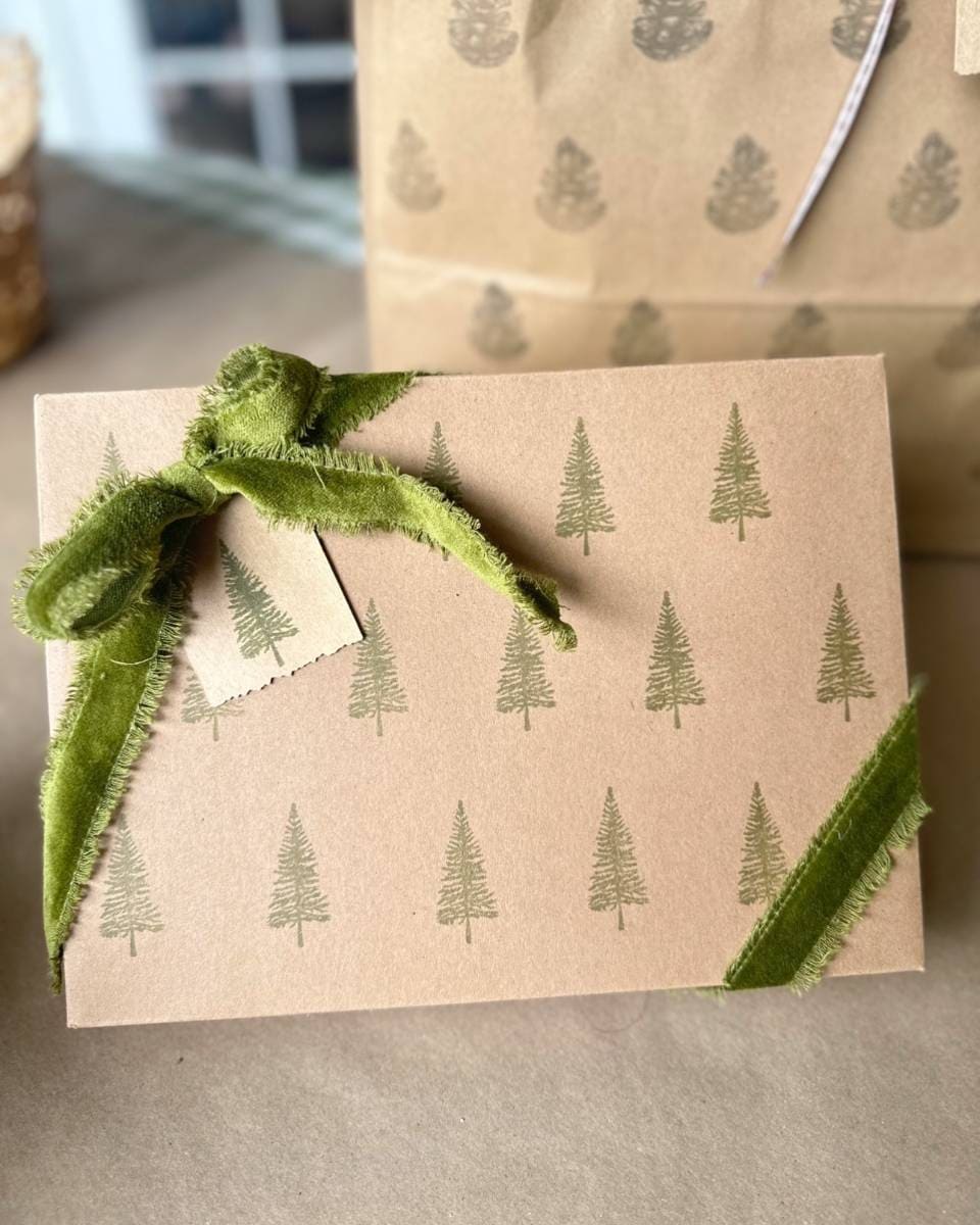 A gift wrapped in brown paper with block print style green tree designs, tied with a green fabric ribbon and a small blank tag attached. In the background, more paper featuring similar tree motifs is partially visible.