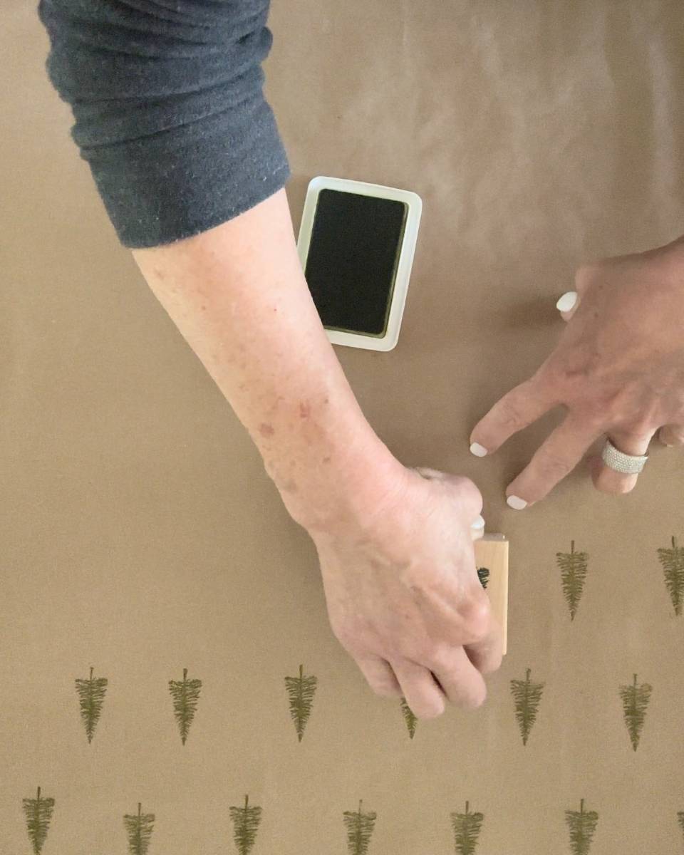 A person crafts block print style gift wrapping paper by stamping small tree designs on brown paper using a rubber stamp and an ink pad.