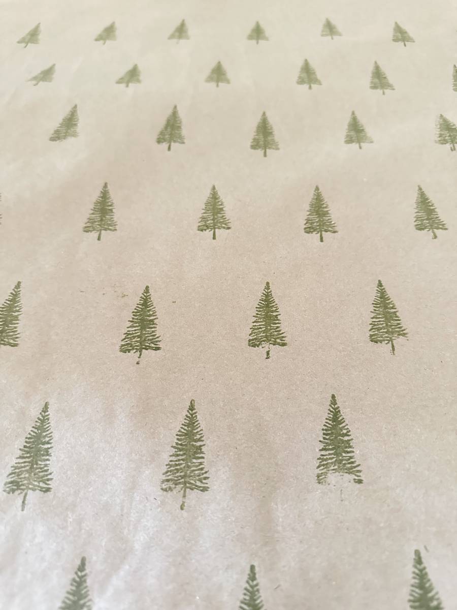 This block print style gift wrapping paper features a sheet of beige with a charming pattern of small green pine trees, evenly spaced in rows.