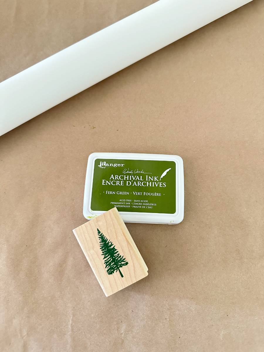 A wooden stamp with a green pine tree design, perfect for block print style gift wrapping paper, sits next to a green archival ink pad labeled "Fern Green" on a brown paper background.