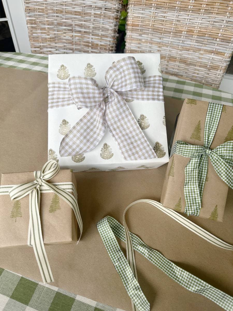 Three gift boxes wrapped in a charming block print style with brown and white tree patterns are tied with plaid and striped ribbons. They rest on a green checkered surface, flanked by two wicker chairs in the background.