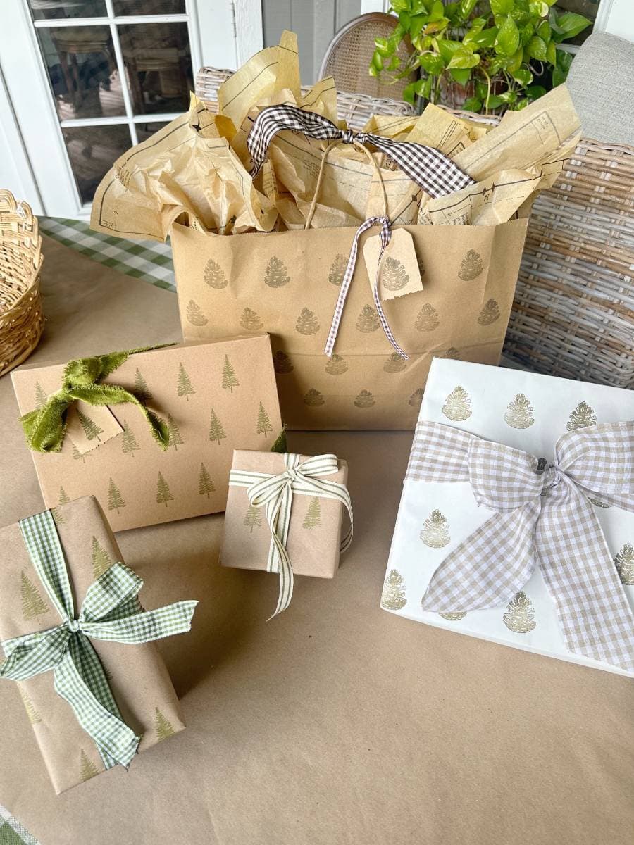 A collection of wrapped gifts on a table features block print style gift wrapping paper. Two packages showcase kraft paper with green tree patterns, another flaunts white paper with a gold pinecone design, all tied with various ribbons. A paper bag with tissue paper stands behind them.