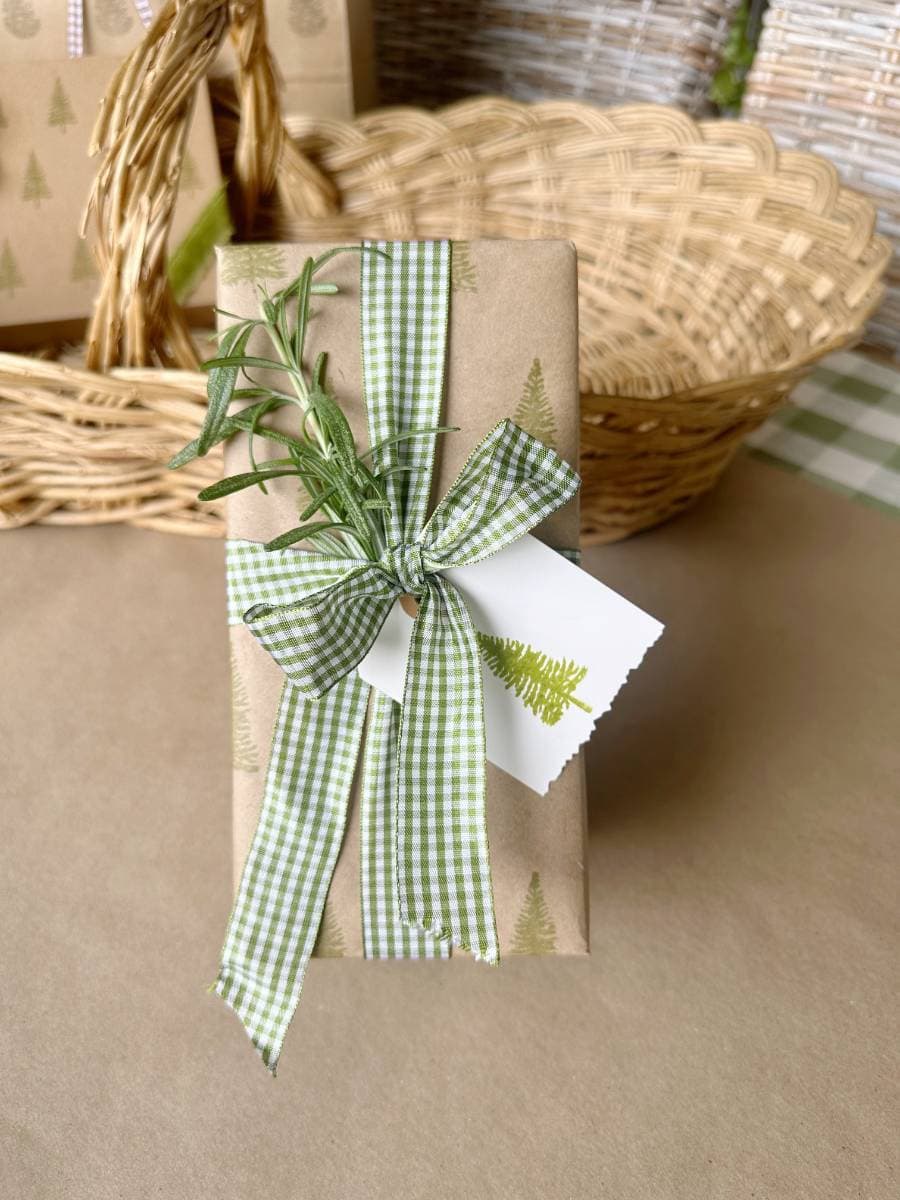 A wrapped gift features tree-patterned block print style paper, adorned with a green gingham ribbon and a sprig of rosemary. A tag with a tree print is attached. The scene is set against a wicker basket and more wrapped gifts, creating an elegant yet rustic appeal.