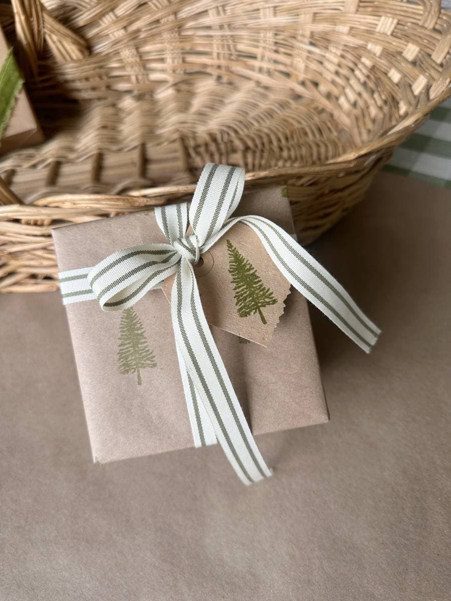 A small gift wrapped in brown paper with a block print style featuring green pine tree stamps, tied with a white and green striped ribbon. A matching tag is attached. The gift is placed in front of a wicker basket.