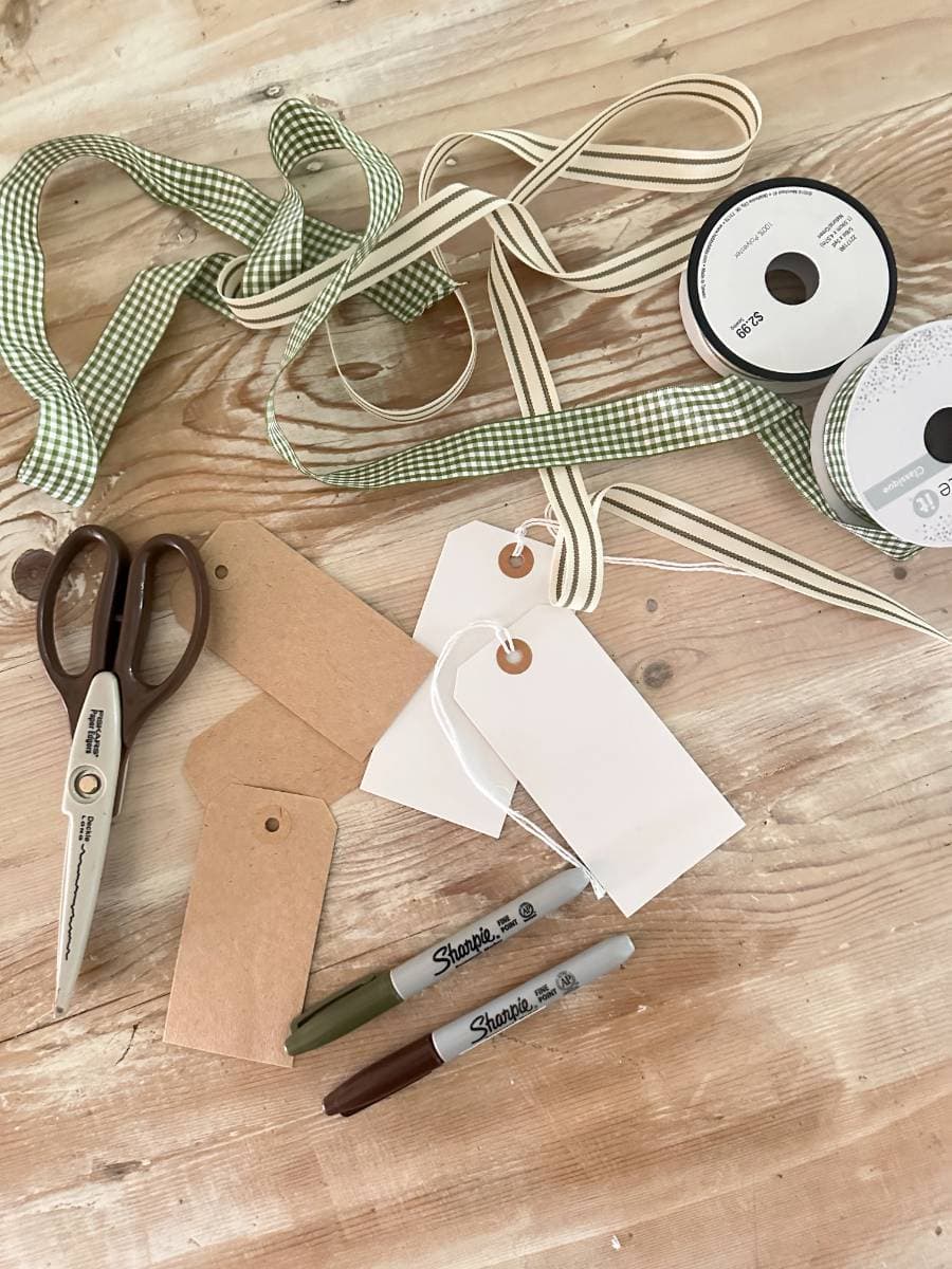 Crafting materials on a table include green and beige ribbons, scissors, brown and white gift tags, two Sharpie markers (one green, one brown), and a roll of block print style gift wrapping paper.