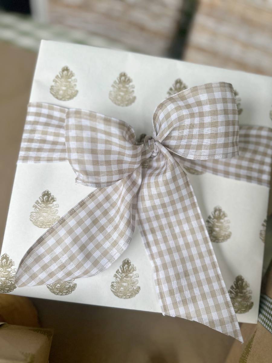 A gift box wrapped in block print style white paper adorned with gold pinecone patterns, tied with a large beige and white checkered ribbon in a bow.