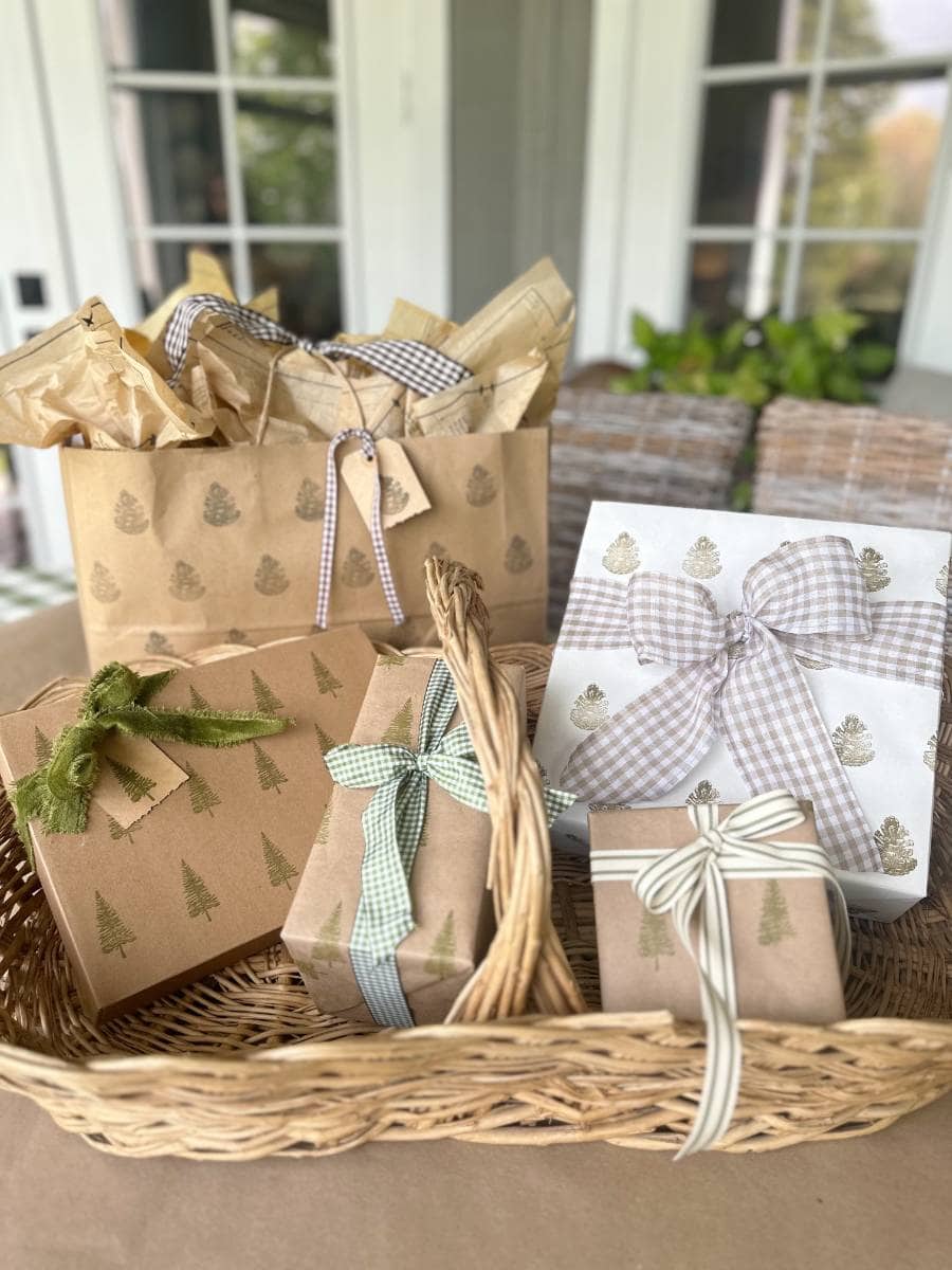 A wicker basket holds several wrapped gifts adorned with block print style gift wrapping paper and bow ribbons. In the background, a gift bag shares similar decorations and tissue paper. The setting is a porch or patio, offering a glimpse of potted plants through the window.