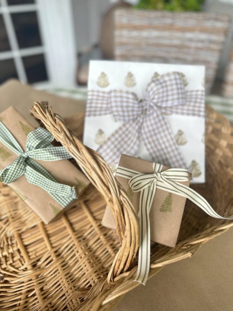 A wicker basket holds three gifts wrapped in brown paper with ribbon bows. Two have green ribbons, while one stands out in block print style gift wrapping paper featuring silver trees on a white background. The scene is cozy and festive.