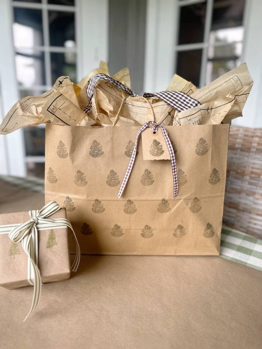 A large gift bag with block print style pine tree designs, filled with tissue paper, has a ribbon and small tag attached. Beside it, a smaller gift box wrapped in matching brown paper with tree prints and ribbon rests elegantly on the table.