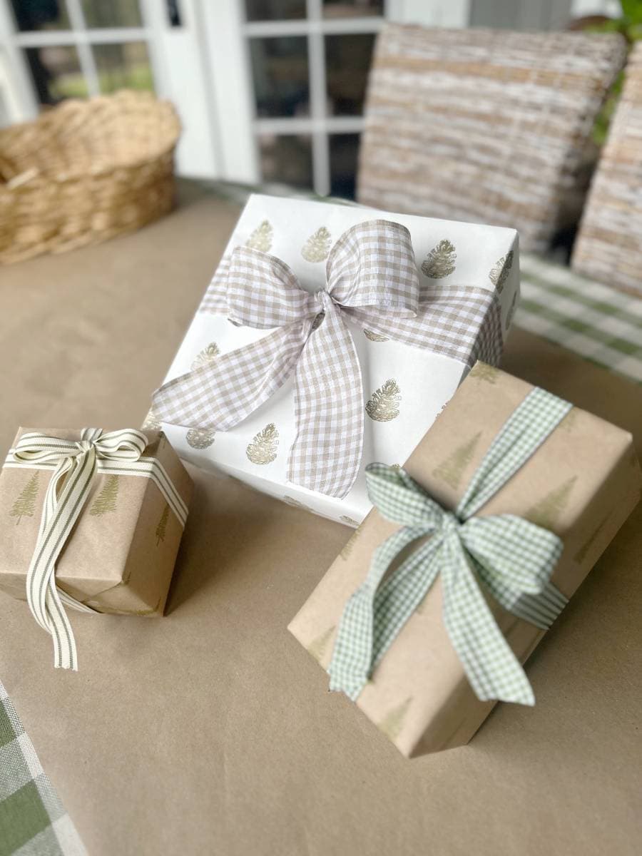 Three wrapped gift boxes with bows are arranged on a table. The largest box features block print style wrapping paper adorned with pine cone patterns and a gingham ribbon. The smaller boxes flaunt tree-printed paper, one with a white ribbon and the other with a green ribbon.