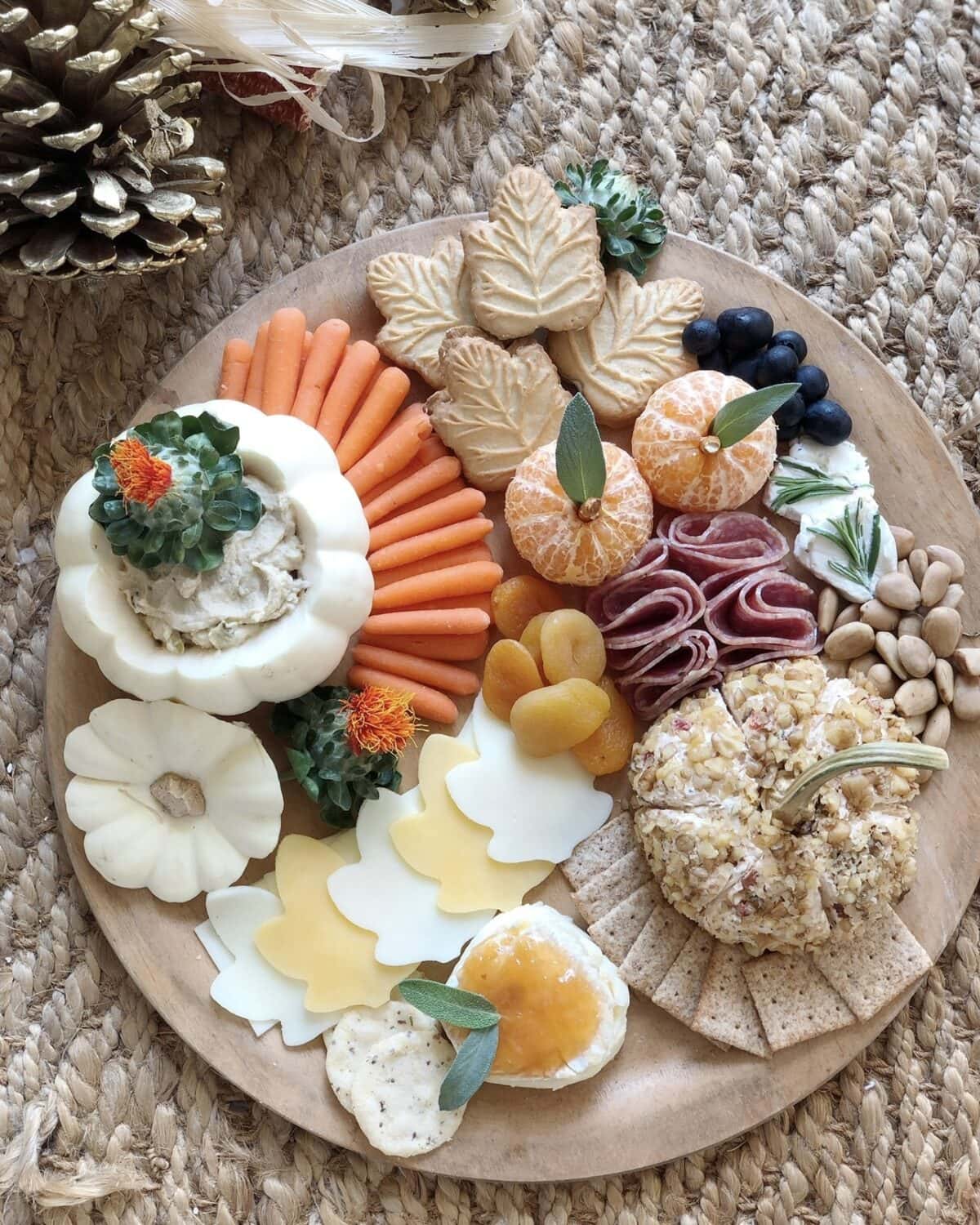 Create a Fun Halloween Snack Board featuring a round wooden board with crackers, carrot sticks, cheese slices, dried apricots, pistachios, blueberries, mandarin oranges, salami, and dips in small white pumpkins for a festive fall display.