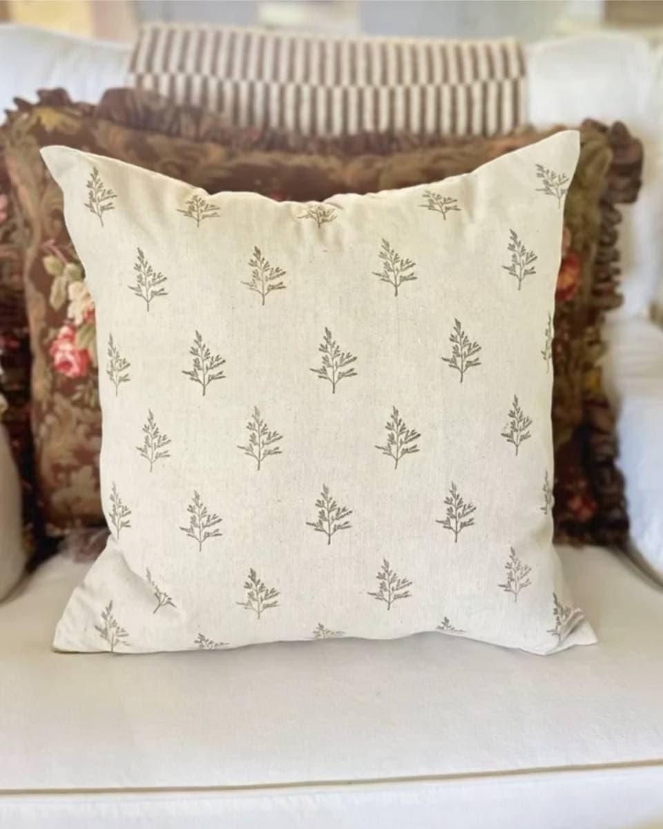 A decorative pillow with an off-white background features small, repeated brown leaf patterns reminiscent of block print style. It's elegantly placed in front of another floral design pillow, set on a white sofa—a perfect accent piece inspired by the charm of artisan gift wrapping paper.