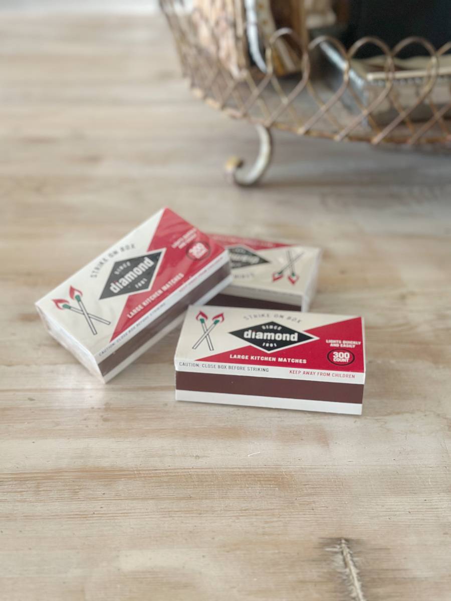 Three boxes of Diamond kitchen matches, ideal for a Decoupage DIY project, sit on a wooden surface. The boxes are primarily red and white with illustrations of crossed matches, while a basket is blurred in the background.