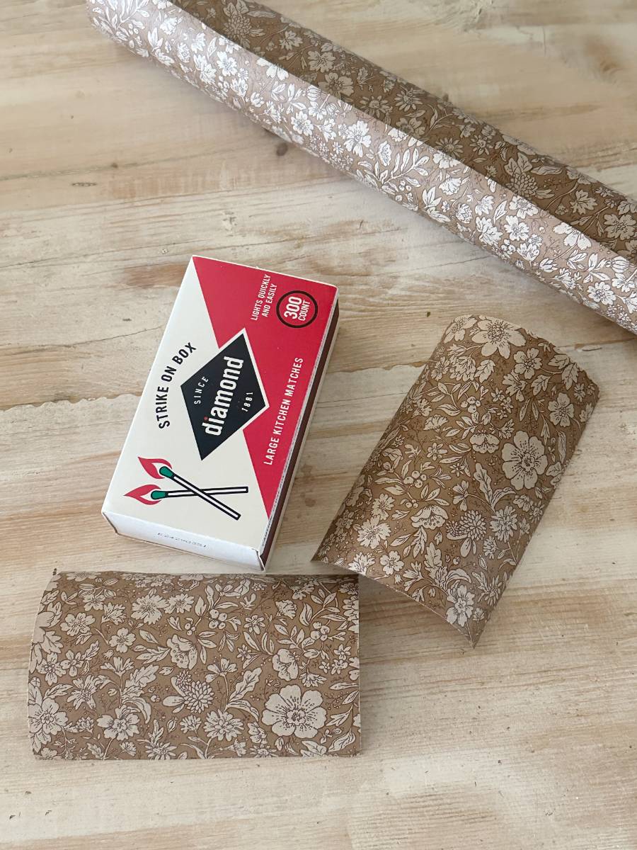 A box of Diamond matches on a wooden surface, alongside sheets of decoupage-decorated paper with a floral pattern. Two pieces are shaped into pillow boxes, and a roll lies in the background, perfect for a crafty DIY project.