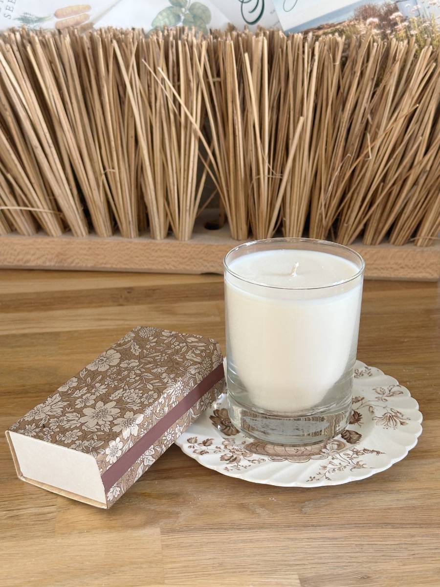 A white candle in a glass sits on a decorative floral-patterned plate. Beside it lies an ornate matchbox, decoupage decorated with a brown and white design. In the background, a decorative arrangement of dried reeds is visible, adding an artsy DIY touch to the scene.