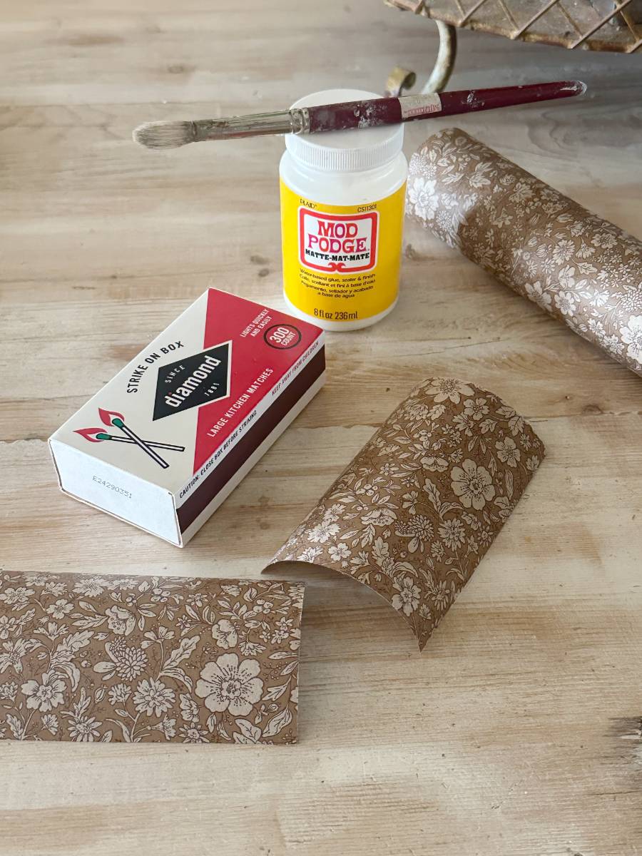 A wooden table showcases a beautifully decoupage-decorated matchbox, resting alongside brown floral paper, a brush, and a jar of Mod Podge adhesive.