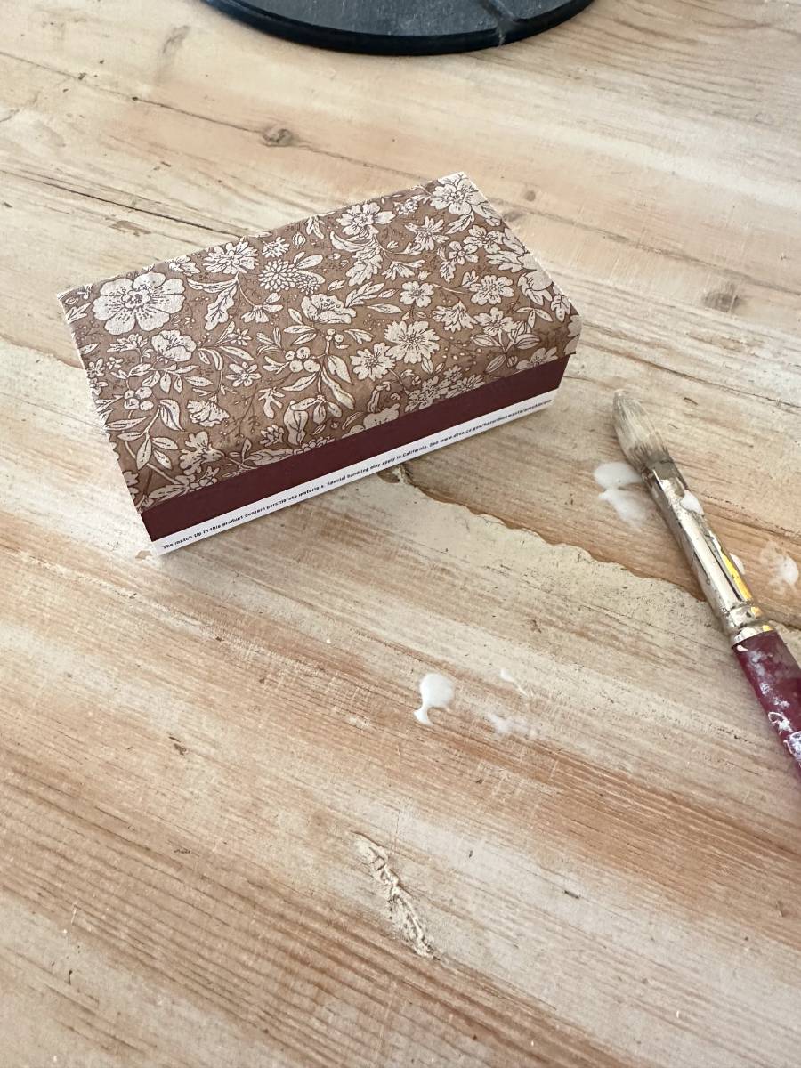 A small, floral-patterned rectangular box sits on a wooden surface with visible wood grain. Nearby, a paintbrush with dried paint hints at a decoupage or DIY project.