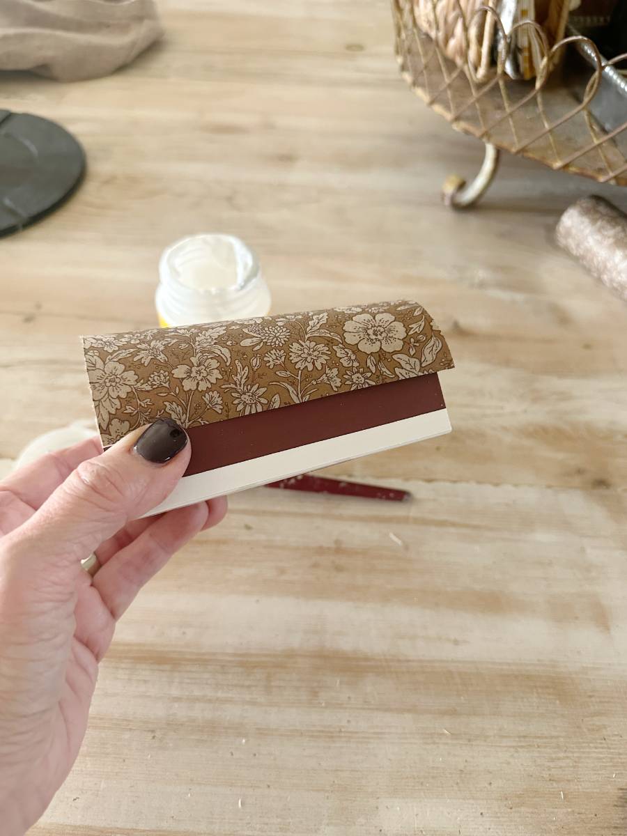 A hand holds a piece of paper folded into three sections, with a floral-patterned top layer and a plain brown middle layer—perfect for decoupage projects. In the background on the wooden table, a jar of white glue and a basket with DIY craft supplies await inspiration.