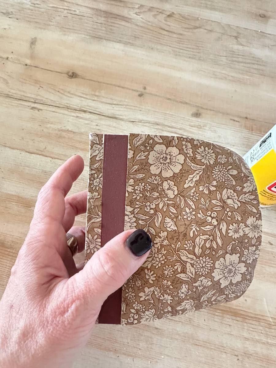 A hand holding a small, decoupage-decorated cardboard pouch against a wooden background. The pouch has a brown strip on one side, and the hand has black nail polish. A yellow container is partially visible on the right.