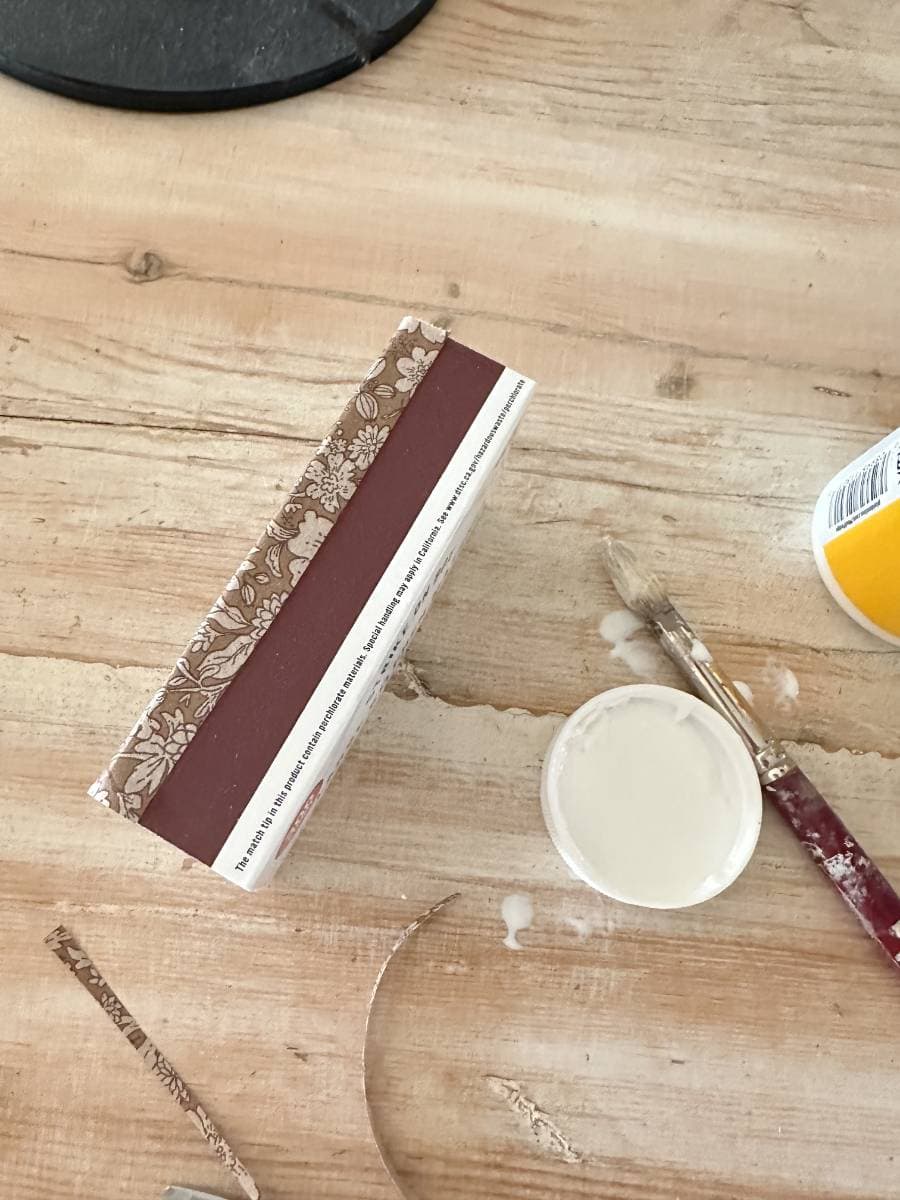 A wooden table features a brown decoupage box, a white paint container with a lid, and a paintbrush dipped in white paint. Scattered pieces of tape or paper strips hint at an ongoing DIY project, perhaps decorating matchboxes for a creative touch.