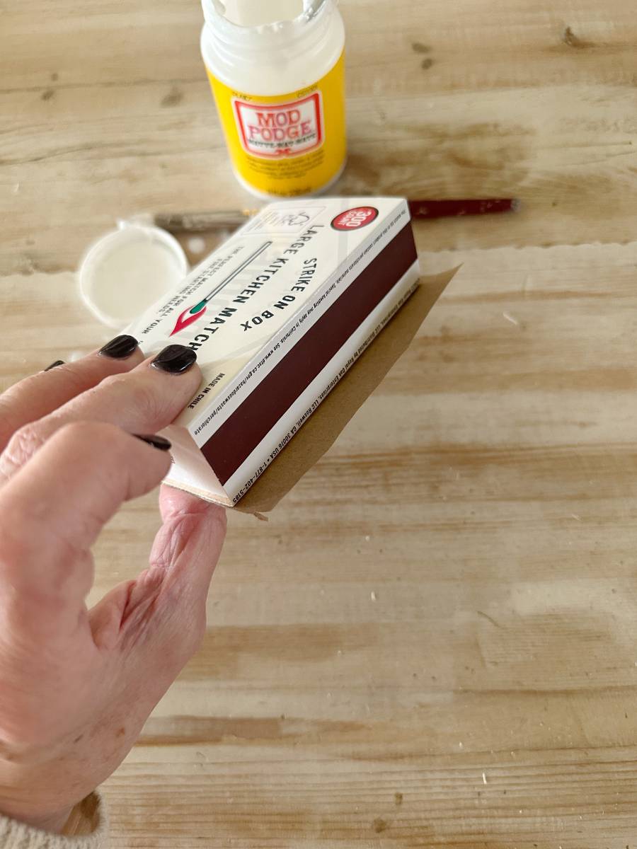 A person with black nail polish applies a Mod Podge sealer to a DIY cardboard box labeled "Campfire Hot Cocoa." Nearby, on the wooden surface, lie a brush and open container of Mod Podge, perfect for decoupage projects or decorated matchboxes.