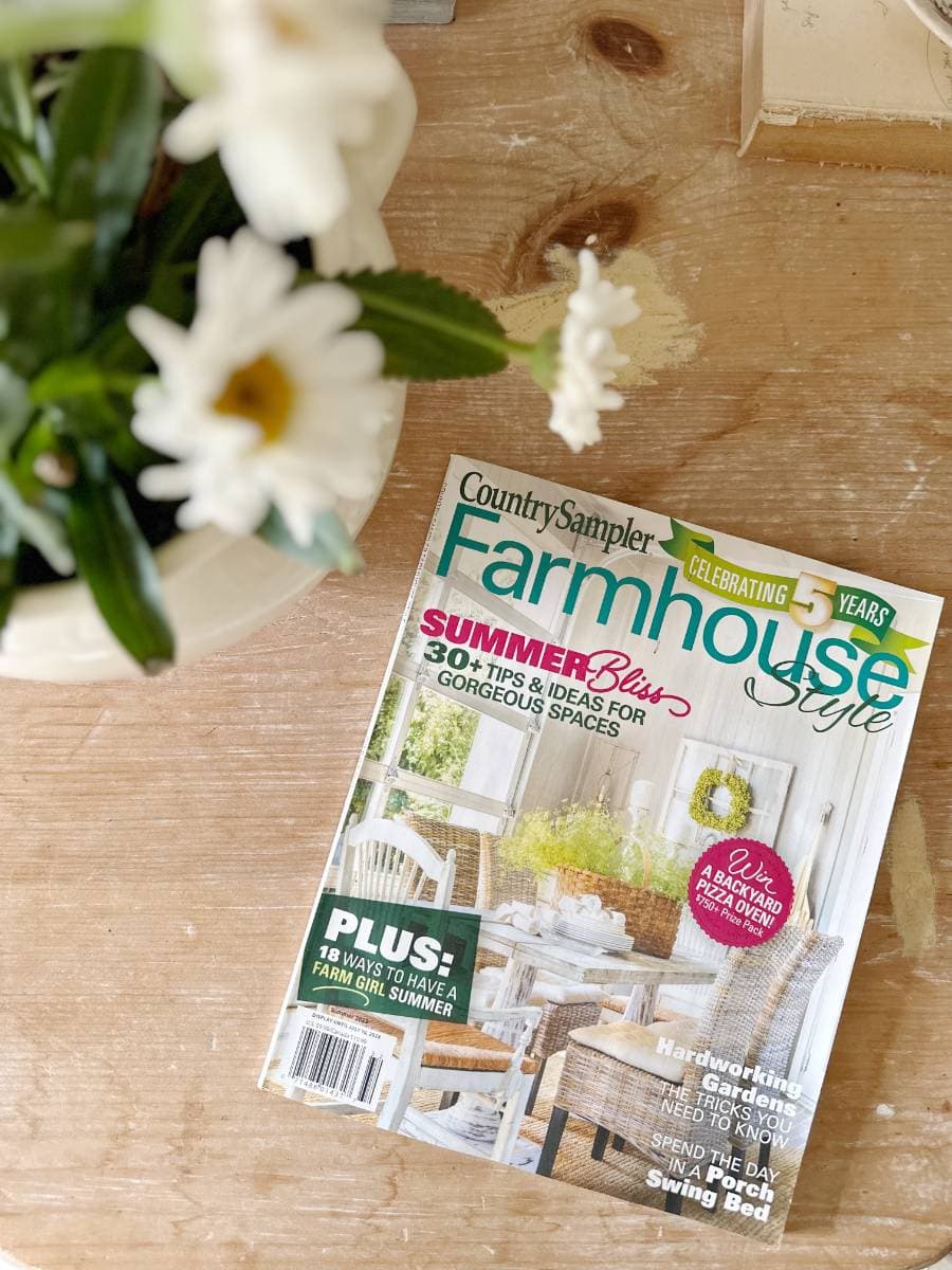 A "Country Sampler Farmhouse Style" magazine sits on a wooden table next to a white daisy plant, featuring summer décor tips, Outdoor Spaces inspiration, and ideas for gorgeous spaces on its cover.