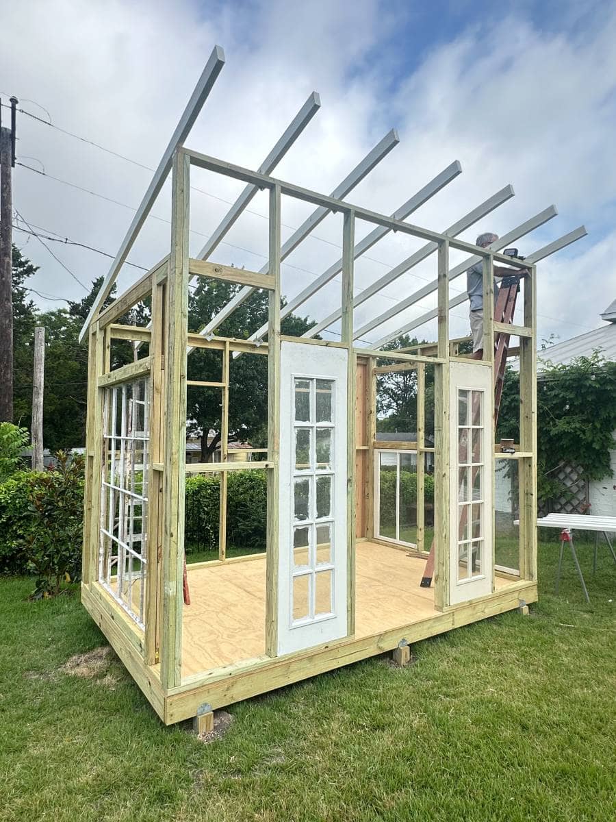 A small wooden structure under construction in a grassy yard, with a wooden frame, multiple glass doors and windows, an unfinished roof, and a person working on a ladder inside—perfect for your next Outdoor Summer Home Tour.