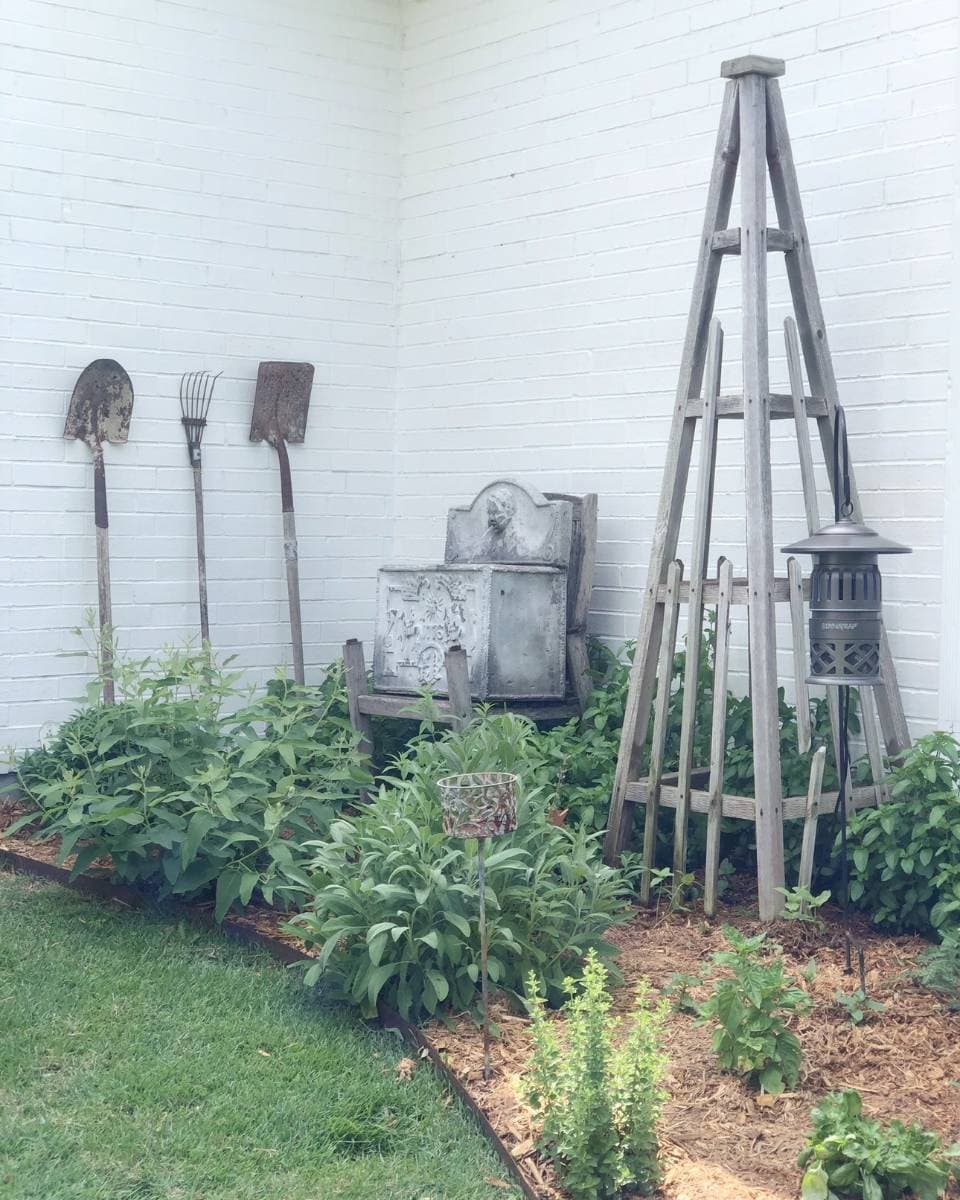A garden corner perfect for an Outdoor Spaces Summer Home Tour, with green plants, old gardening tools on a white brick wall, a rustic bench, trellis, lantern, and bird feeder nestled among mulch and lawn.