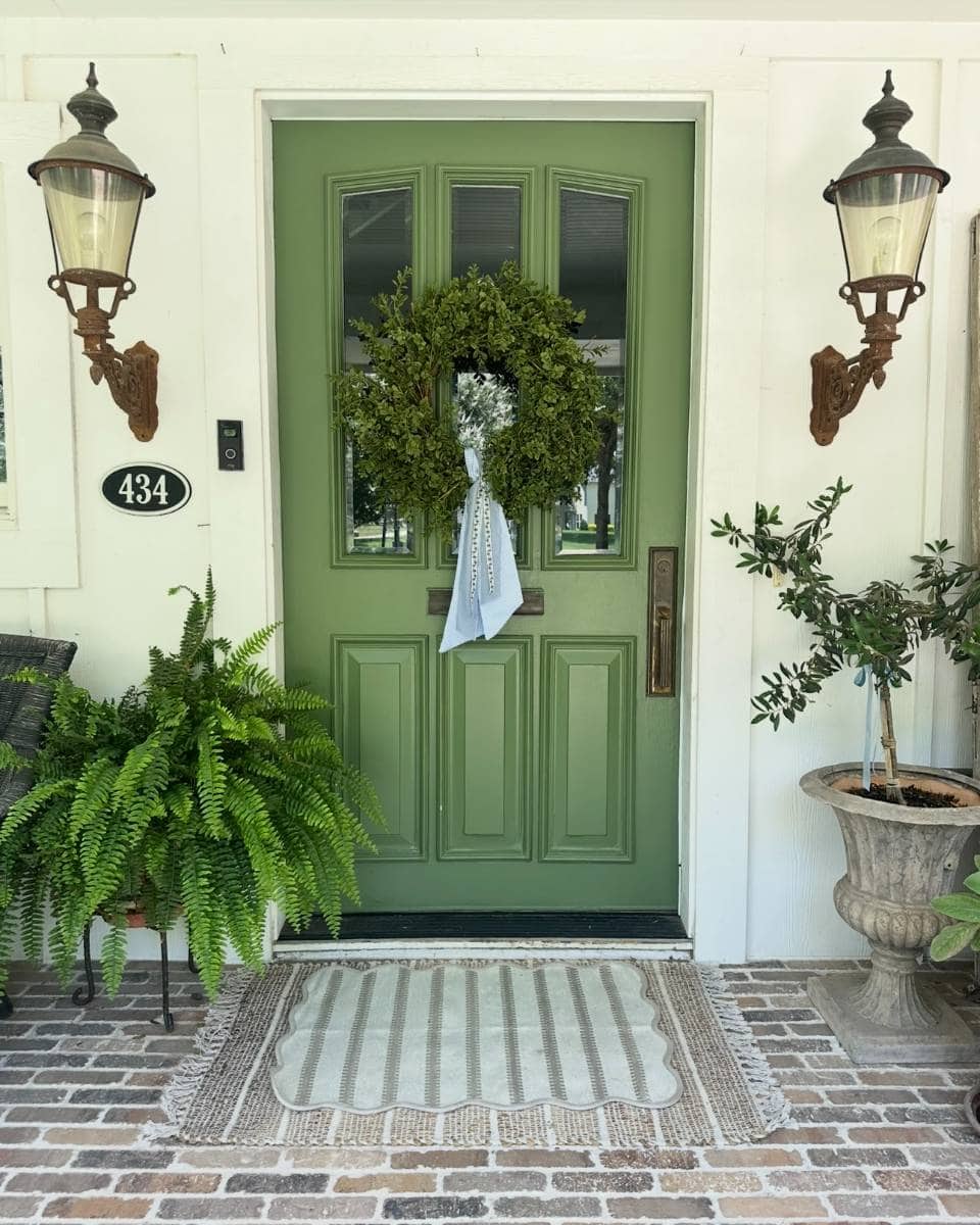 A green front door with a wreath and white ribbon, flanked by lanterns, welcomes you to this charming Outdoor Spaces Summer Home Tour. Potted plants and a striped doormat enhance the brick porch, while house number 434 is proudly displayed.