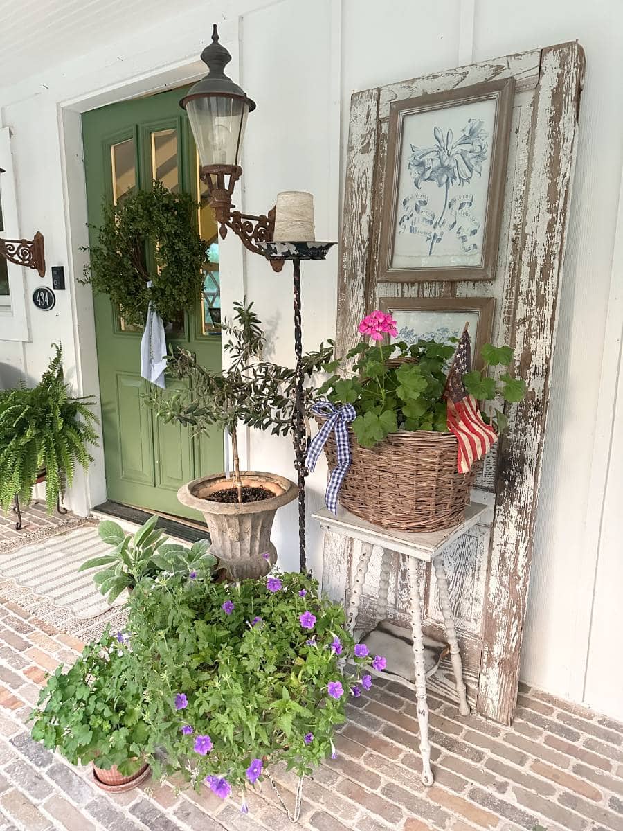 A rustic porch perfect for an Outdoor Spaces Summer Home Tour, featuring a green door, wall lamp, potted plants, a wicker basket with flowers and American flag, vintage botanical art in a wooden frame, and a striped rug on the brick floor.