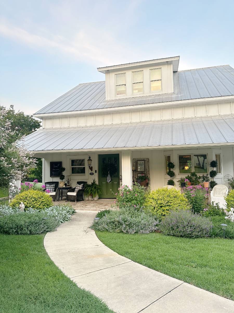 A white farmhouse with a metal roof, front porch, potted plants, and rocking chairs showcases inviting outdoor spaces and lush landscaping along a curved sidewalk under a blue sky—perfect for a Summer Home Tour.