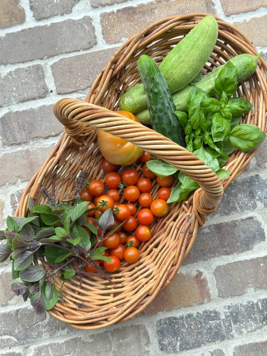 A wicker basket filled with cherry tomatoes, a large yellow tomato, cucumbers, fresh basil, and purple basil rests on a brick surface—perfect for an Outdoor Spaces Summer Home Tour display.