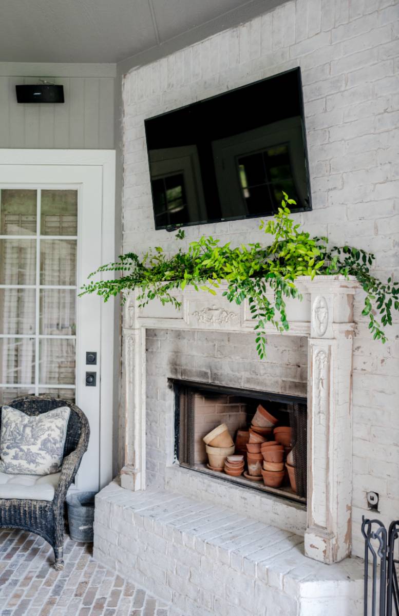 A white brick fireplace with stacked terracotta pots inside, green foliage on the mantel, and a TV above creates cozy charm. To the left, a wicker chair with a patterned cushion welcomes you—perfect for an Outdoor Spaces Summer Home Tour.