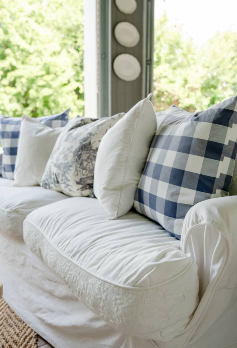 A white sofa with patterned and plain throw pillows, including blue and white checkered designs, sits by a window overlooking greenery—a perfect spot for relaxing during an Outdoor Spaces Summer Home Tour. Decorative plates hang on the nearby wall.