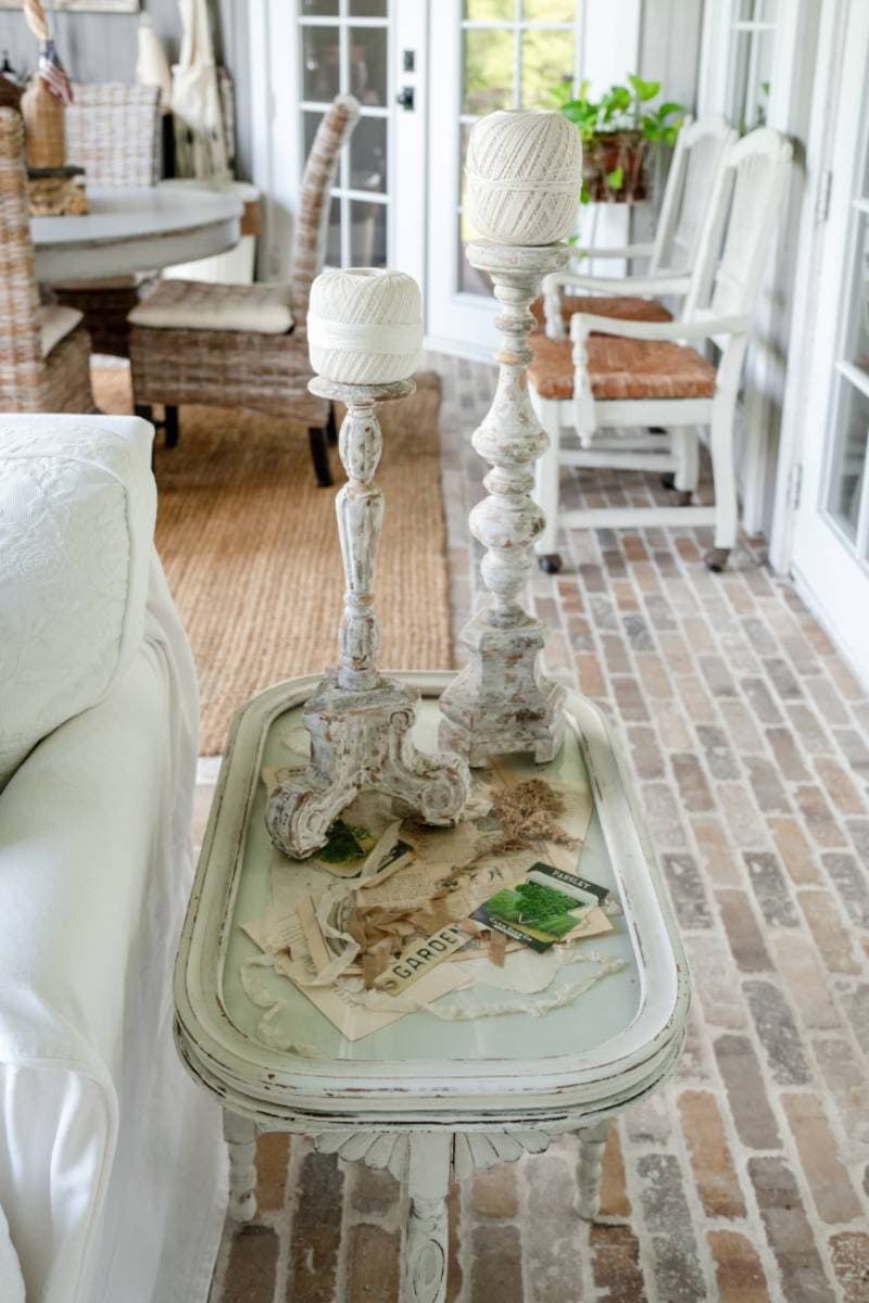 A small vintage side table with a distressed white finish holds two twine-wrapped candlesticks and seed packets—perfect inspiration for your Outdoor Spaces Summer Home Tour. The table rests on a brick floor beneath sunlit wicker and white-painted furniture.