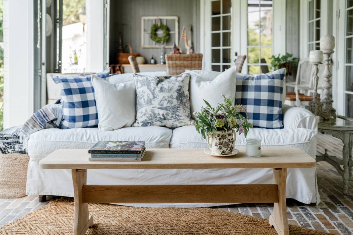 A cozy living room with a white sofa adorned with blue and white patterned pillows, a wooden coffee table holding books, a floral vase, and a candle sits on a woven rug near glass doors—perfect for an Outdoor Spaces Summer Home Tour.