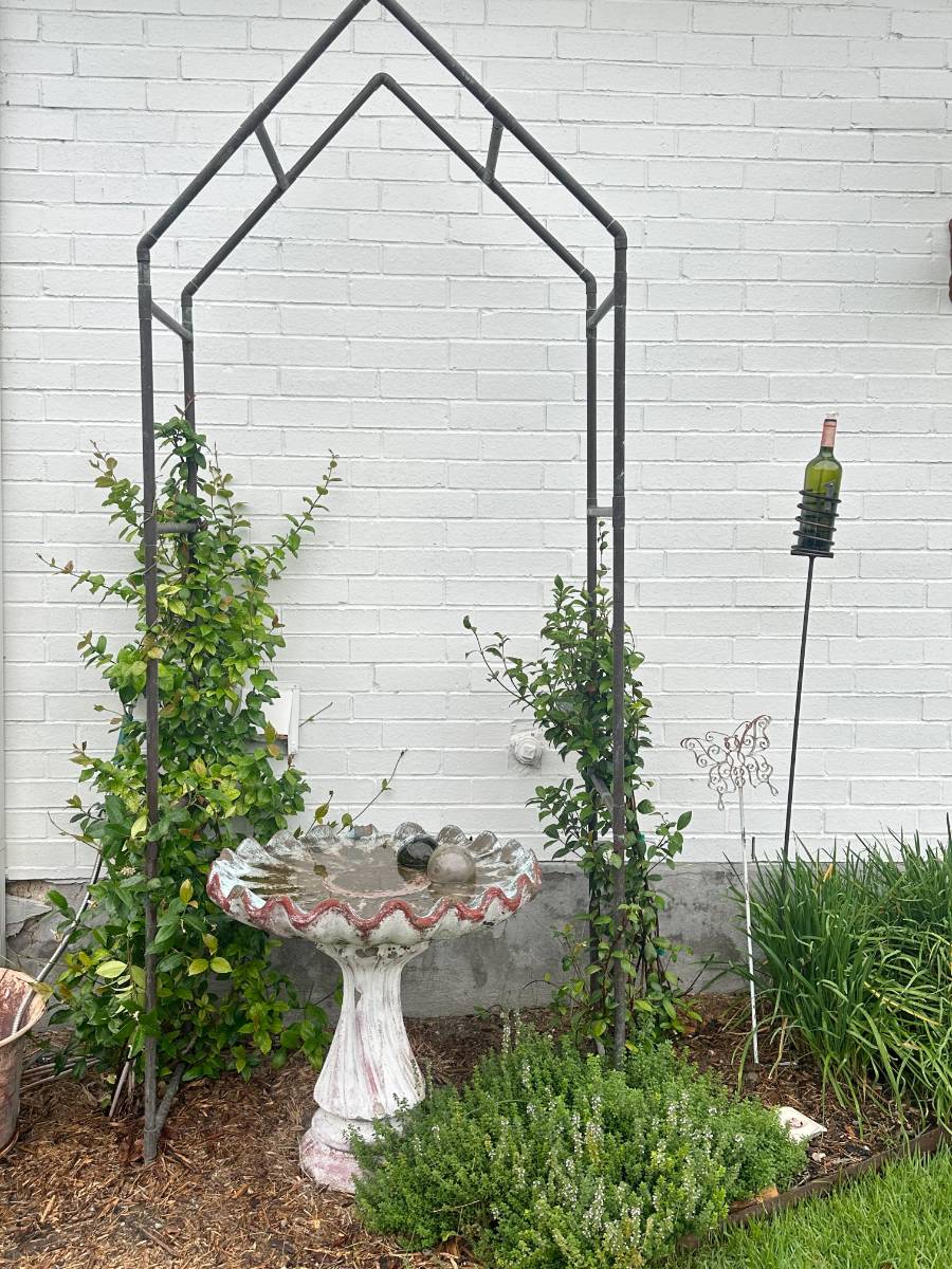 A decorative metal arch stands over a weathered birdbath with red trim, surrounded by green plants against a white brick wall. Perfect for an Outdoor Spaces Summer Home Tour, a wine bottle rests on a metal holder beside the birdbath.