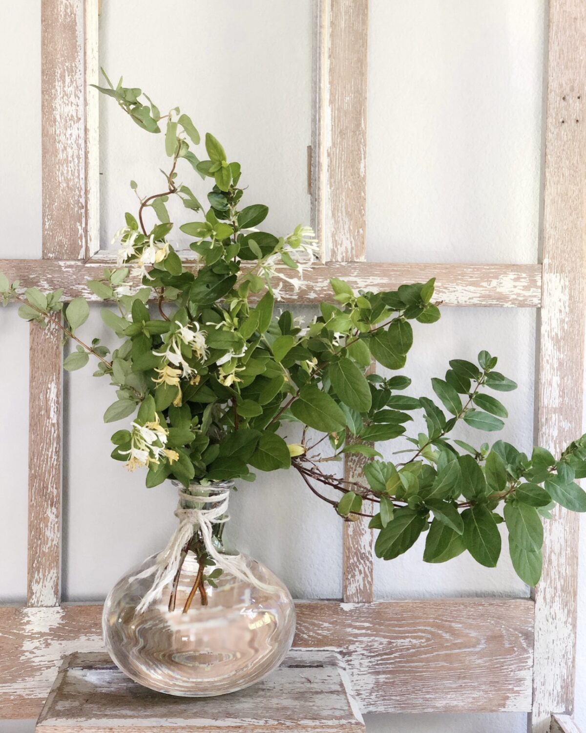 A glass vase filled with green leafy branches and small white flowers sits on a rustic wooden shelf, adding fresh charm to your summer home tour in front of a distressed, light-colored wooden window frame.