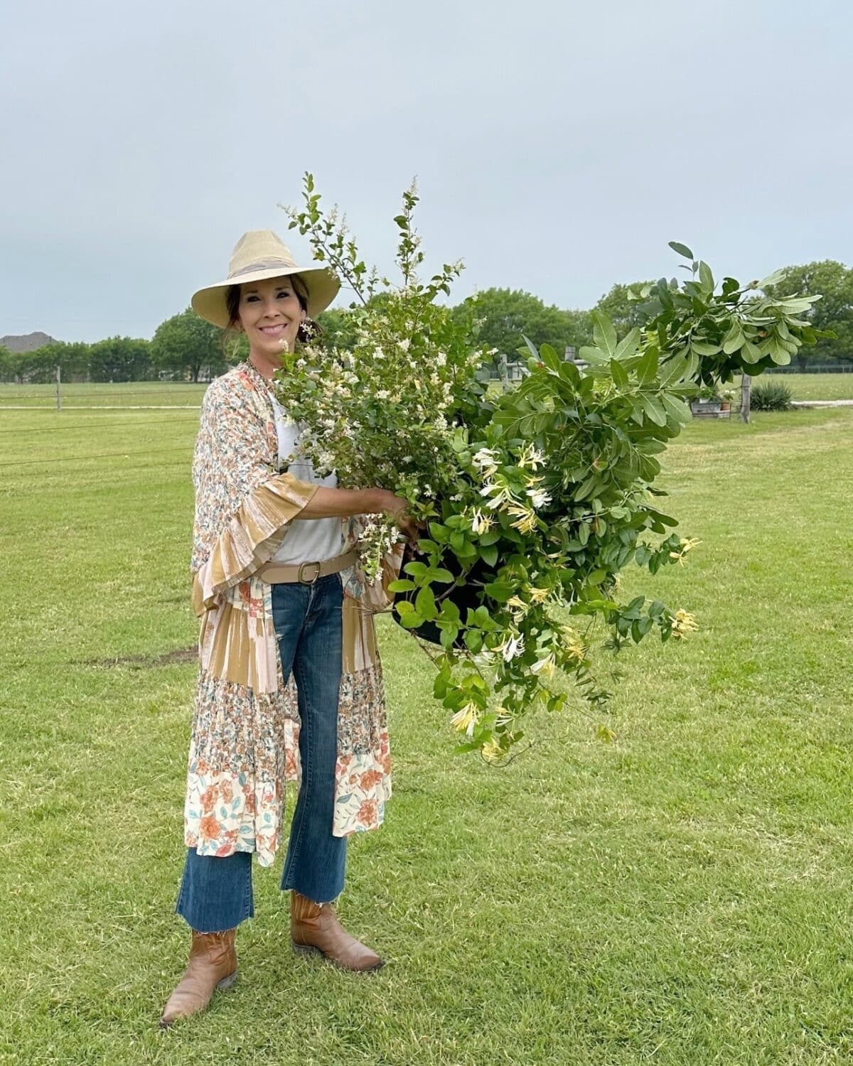 A smiling woman wearing a hat, kimono, jeans, and boots stands on grass holding a large bundle of leafy branches and flowers—an inviting scene perfect for any Outdoor Spaces Summer Home Tour. Green trees and a cloudy sky complete the backdrop.
