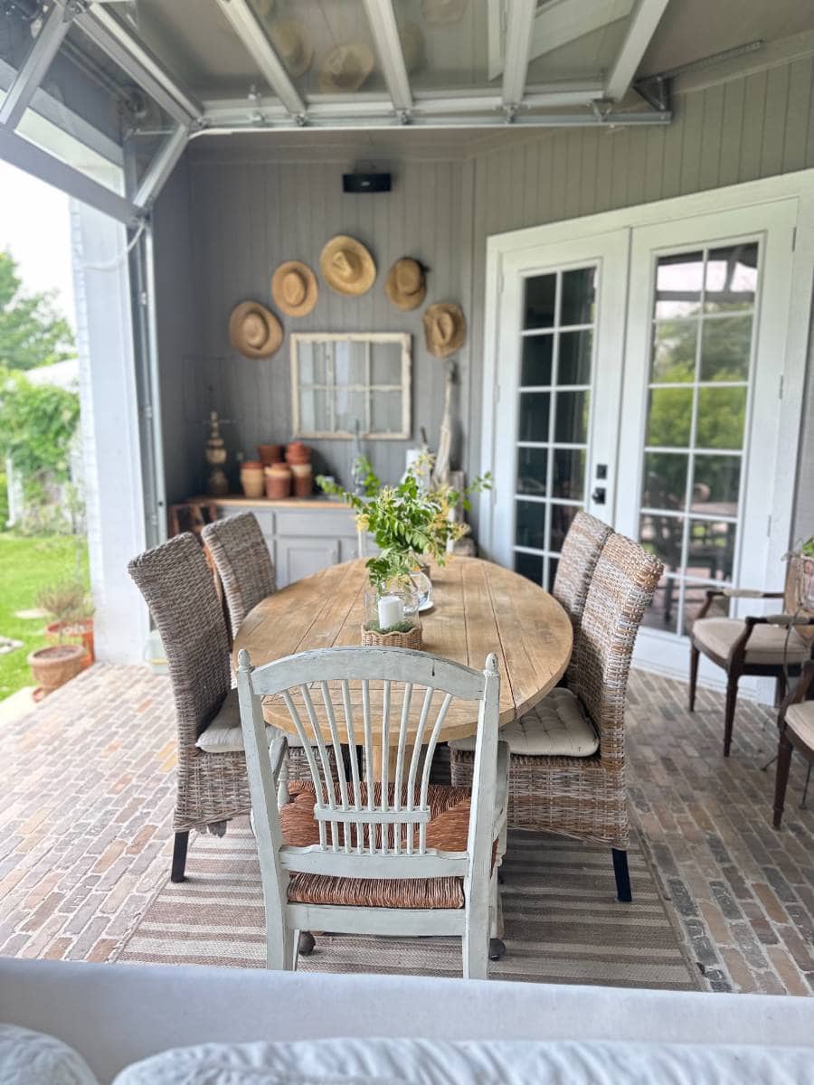 A cozy patio from an Outdoor Summer Home Tour features a wooden oval dining table surrounded by wicker chairs on a striped rug. Woven hats hang on a gray wall above shelves with potted plants, while French doors open to the green yard.