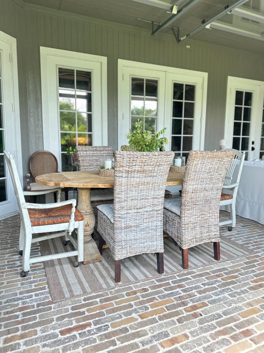 A round wooden table with mixed chairs, including wicker and painted styles, sits on a striped rug on a brick patio for an inviting Outdoor Summer Home Tour. A potted plant decorates the center of the table in front of three white French doors.