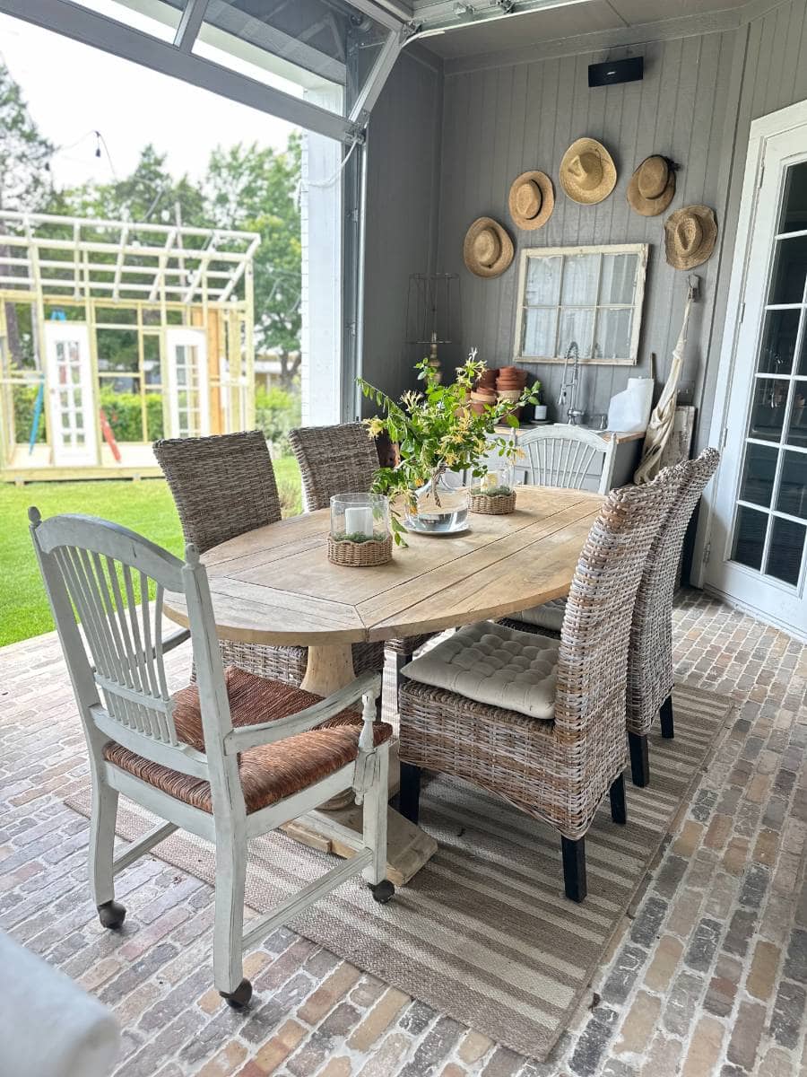 A cozy outdoor dining area with a wooden table, wicker and cushioned chairs, a striped rug, potted plants, and hats hanging on a gray wall—perfect for an Outdoor Summer Home Tour. A play structure and lush greenery are visible through open doors.